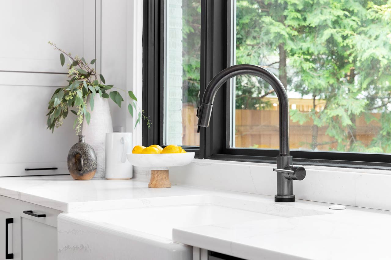 Kitchen sink detail shot in a modern, renovated kitchen with black window frames, a dark faucet, white cabinets, farmhouse sink, and cozy decor