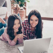 Mother and her little curious daughter are lying on a floor at home and using a laptop