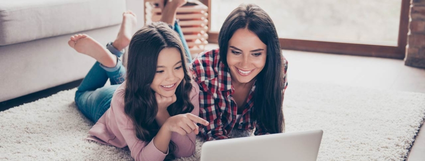 Mother and her little curious daughter are lying on a floor at home and using a laptop
