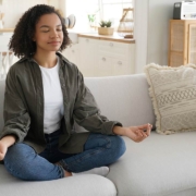 A serene Black woman meditates on a couch, eyes closed, in a sunlit, stylish living room